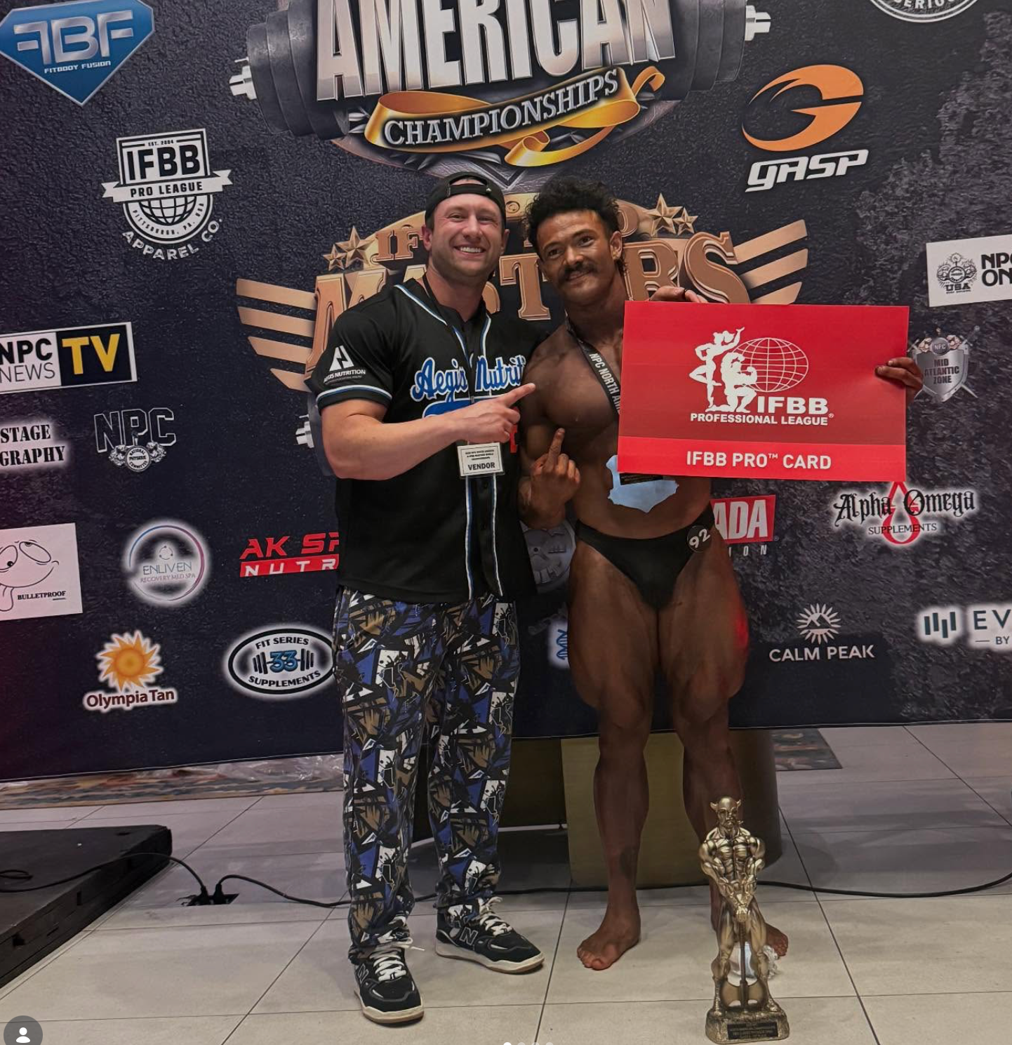 Bodybuilder holding a trophy and card, posing with a man in front of a backdrop.