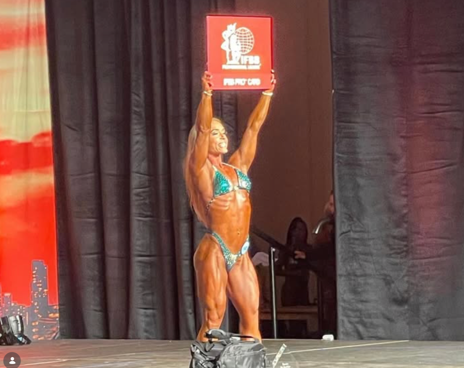 Female bodybuilder, stage, holding up a sign. She's wearing a teal bikini, muscles flexed, dark backdrop.