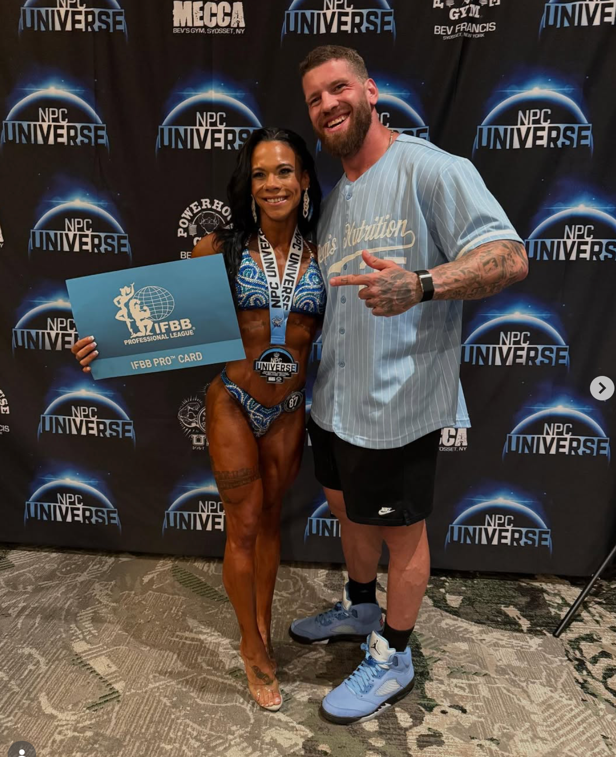 Bodybuilder in patterned bikini holding award, posing with a man in front of a backdrop with logos.