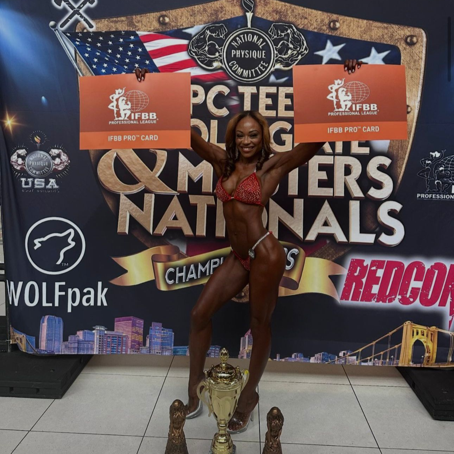 Bodybuilder holding awards and trophy on stage, posing in red bikini.