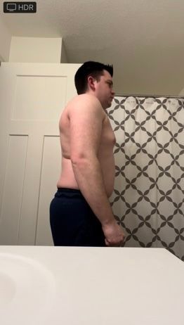 Man in a bathroom, facing sideways, shirtless and wearing dark sweatpants, looking toward the right.
