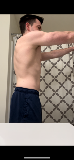 A person, shirtless, stretches arms horizontally, standing in a bathroom. Navy blue pants. Back is visible.