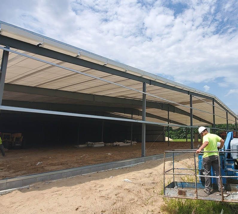 Construction worker on a lift near a large steel-framed building with a white roof and dirt floor.