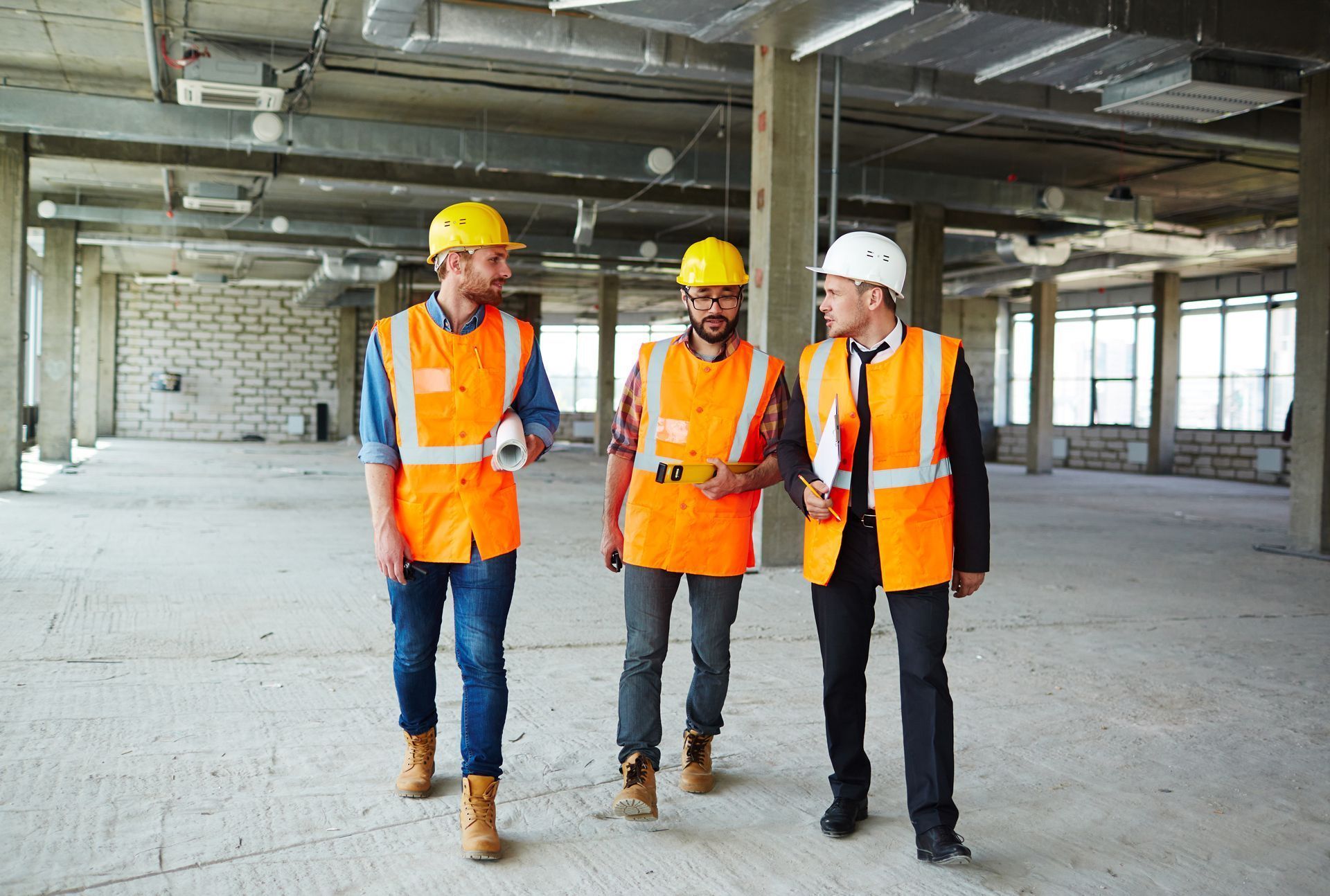 Three construction workers in hard hats and vests on a construction site; one holds blueprints.