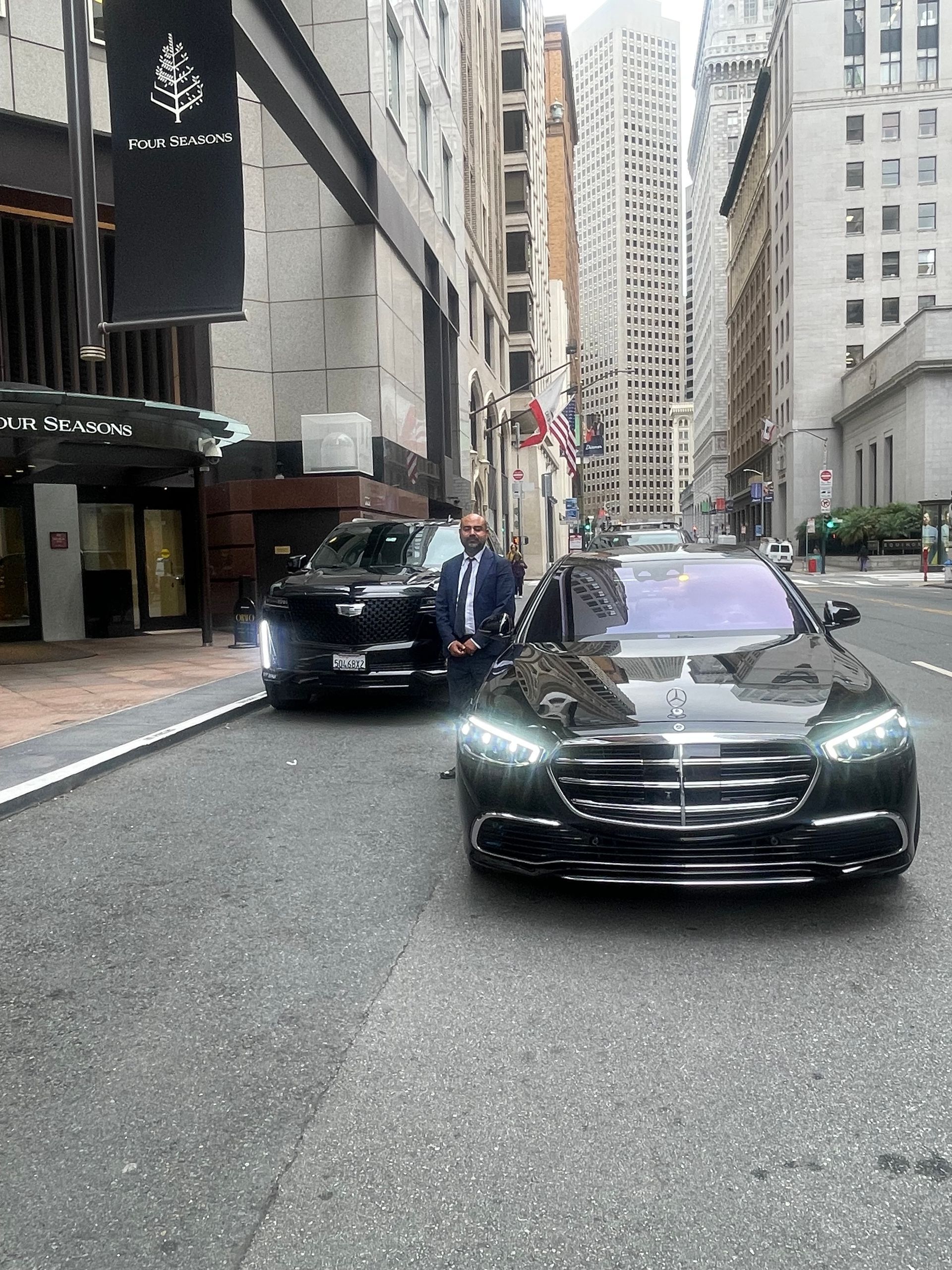 A person in a suit stands between a black sedan and an SUV parked on a city street near a tall building.