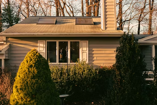 Tan house with a window, white shutters, and bushes. Chimney and skylights on roof.