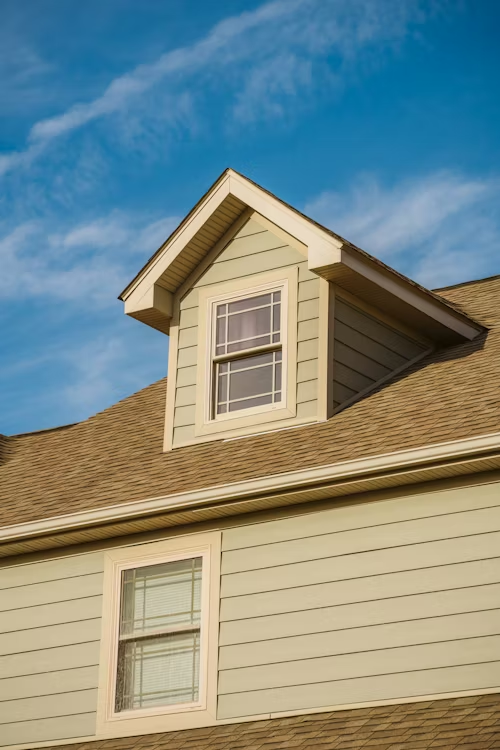 A light green gabled dormer window on a brown shingled roof against a bright blue sky.
