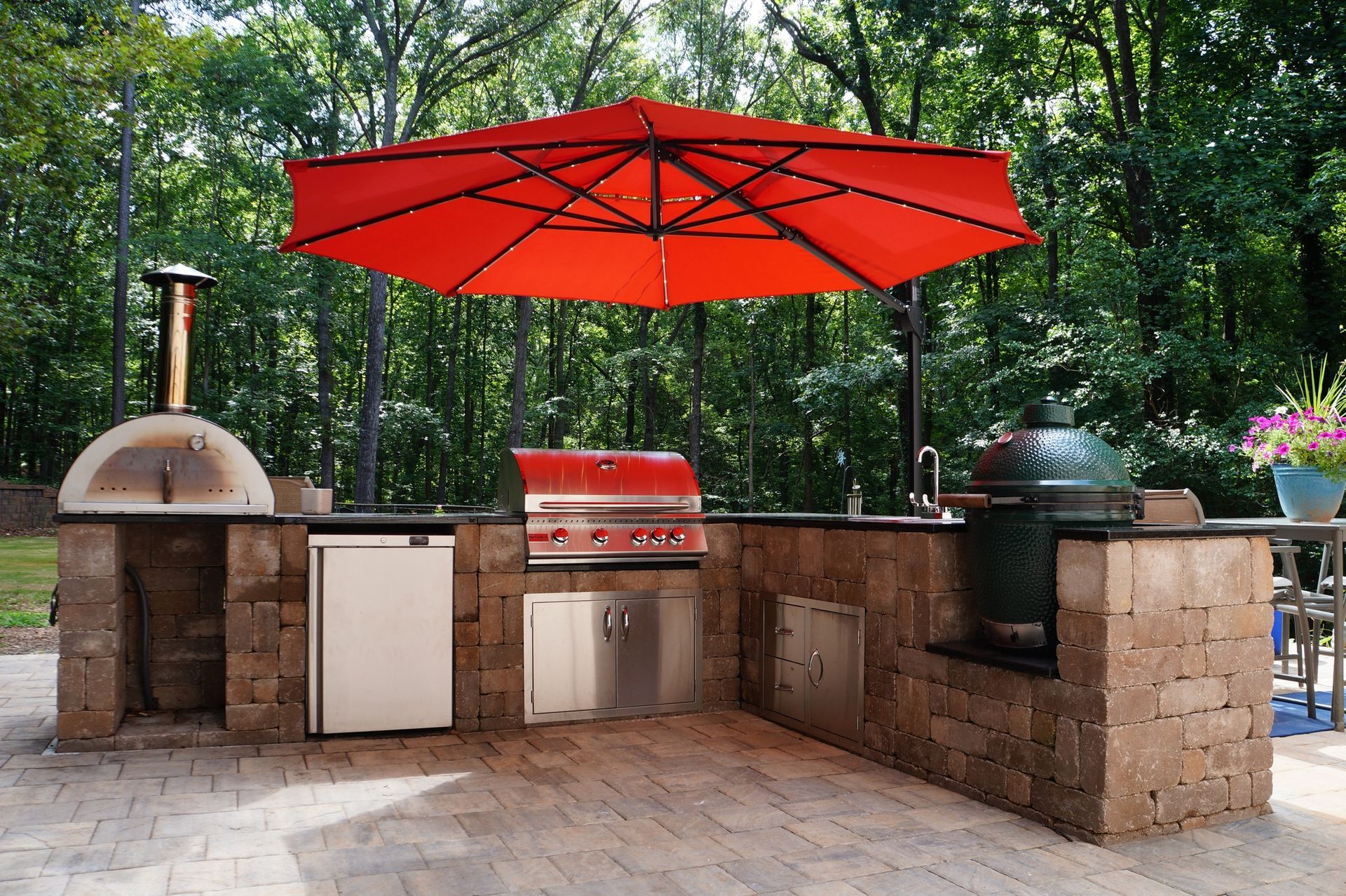 Outdoor brick grill station with red umbrellas, pizza oven, and barbecue grills in a leafy backyard