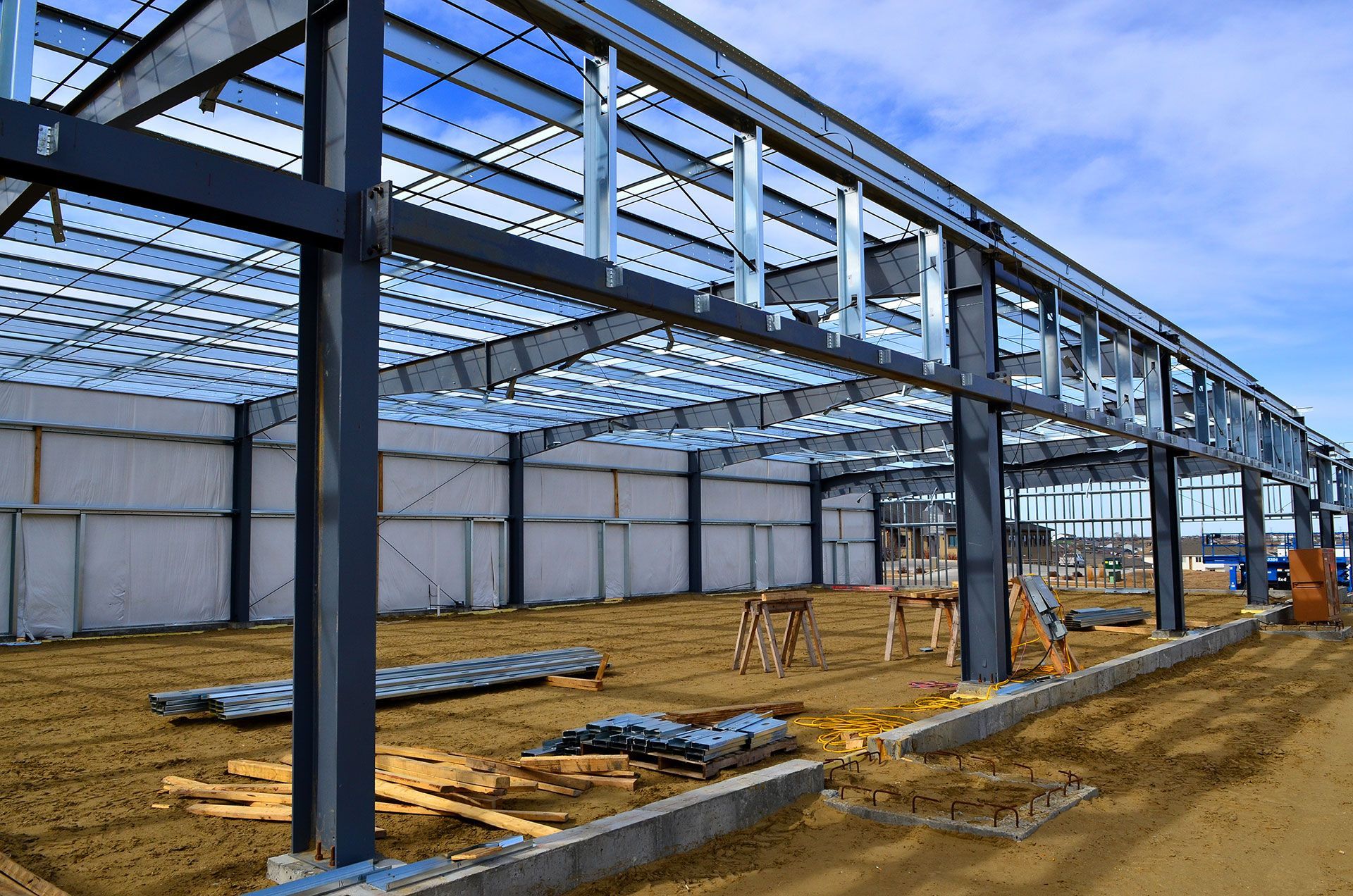 Steel-framed warehouse under construction with concrete foundation and scattered materials on dirt ground.