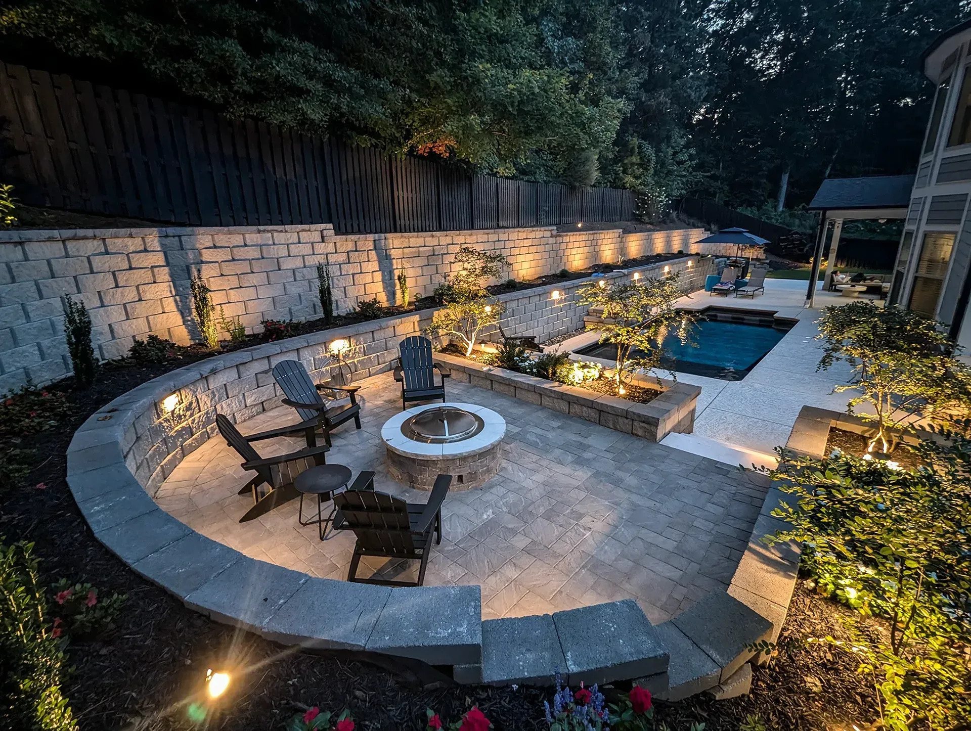 Modern outdoor patio with curved stone seating, fire pit, pool, and ambient garden lighting