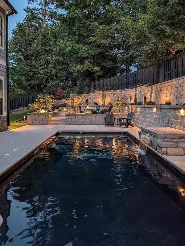 Narrow backyard pool at dusk with stone patio, warm lights, and lush trees along a retaining wall