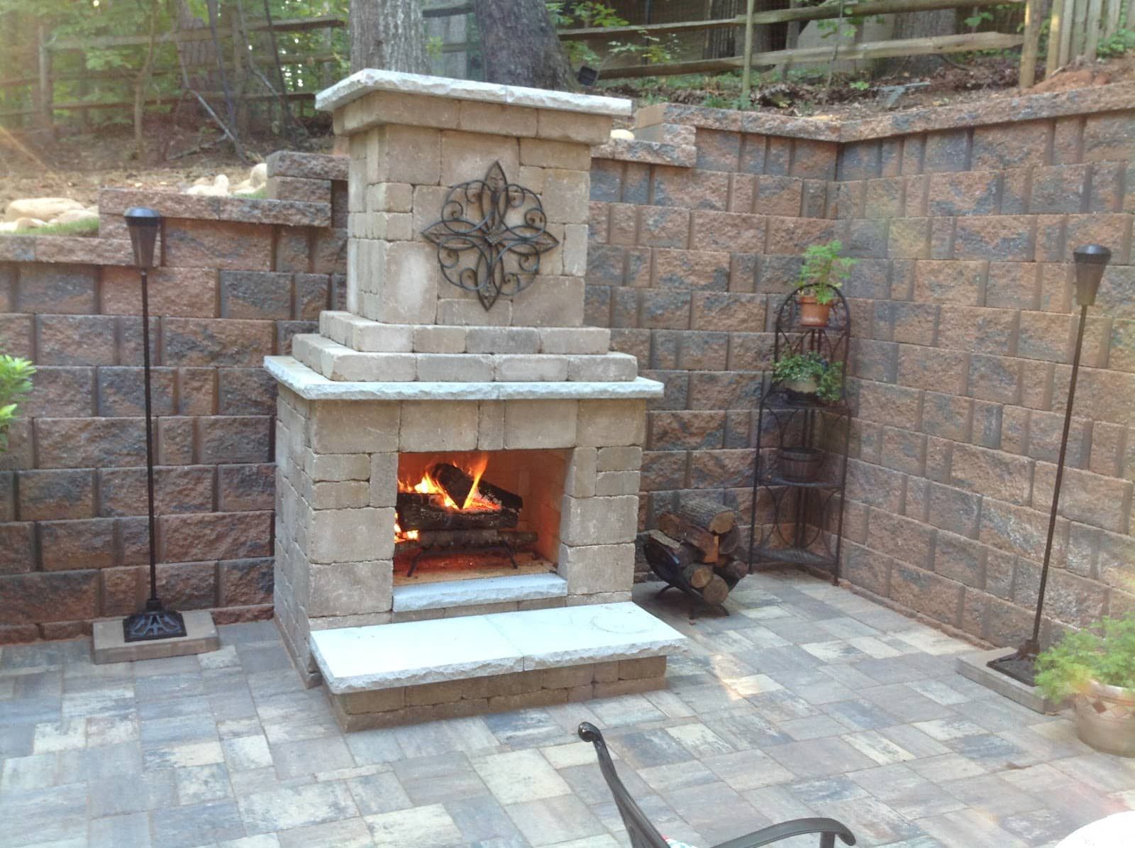 Stone outdoor fireplace burning in a brick patio with retaining walls and potted plants