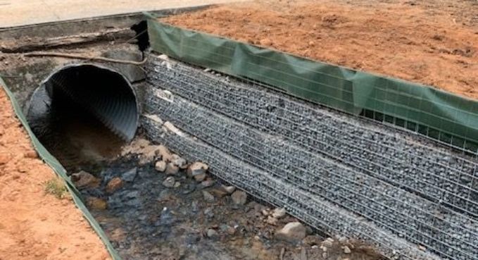 Culvert pipe outlet with rock-filled gabion retaining wall on a dirt slope.