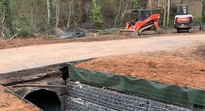 Road construction site with excavator and dump truck beside a damaged culvert and erosion control tarp