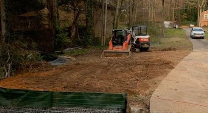 Two construction vehicles grading a dirt area beside a road and wooded path
