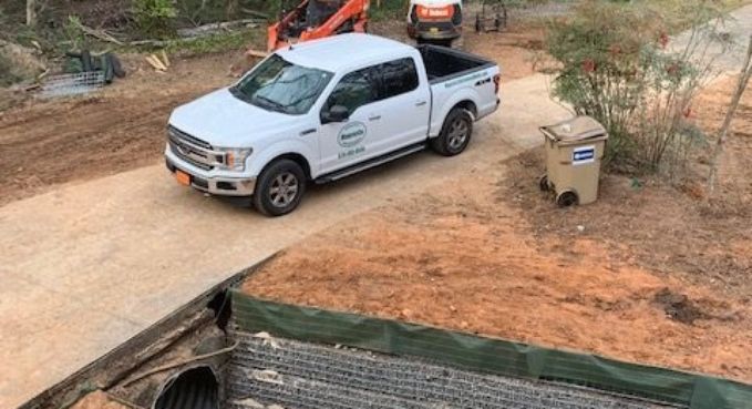 White pickup truck parked on a dirt road beside a drainage ditch and trash bin under construction barriers