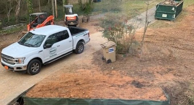 White pickup truck parked on dirt near a trash bin, orange tractor, and green dumpster in a yard