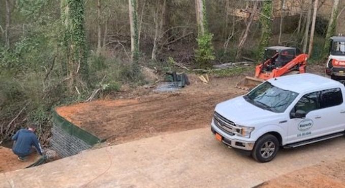 Construction site with white pickup, orange excavator, and worker clearing a dirt slope by the woods