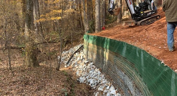Green tarp-lined retaining wall beside wooded slope, with construction equipment and worker on a dirt path