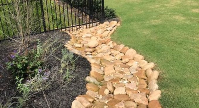Rock-lined garden path beside green lawn and mulch, leading to a black metal gate.