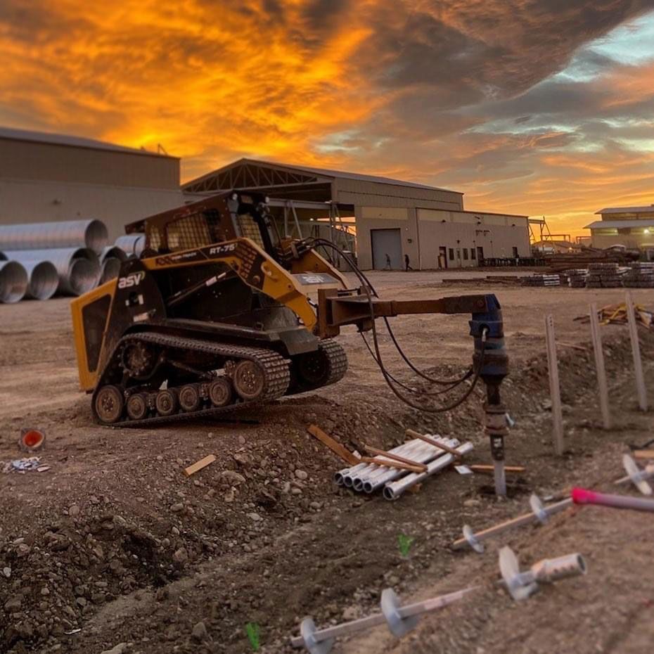 Small tracked construction machine at a worksite at sunset, with industrial buildings and orange clouds in the background