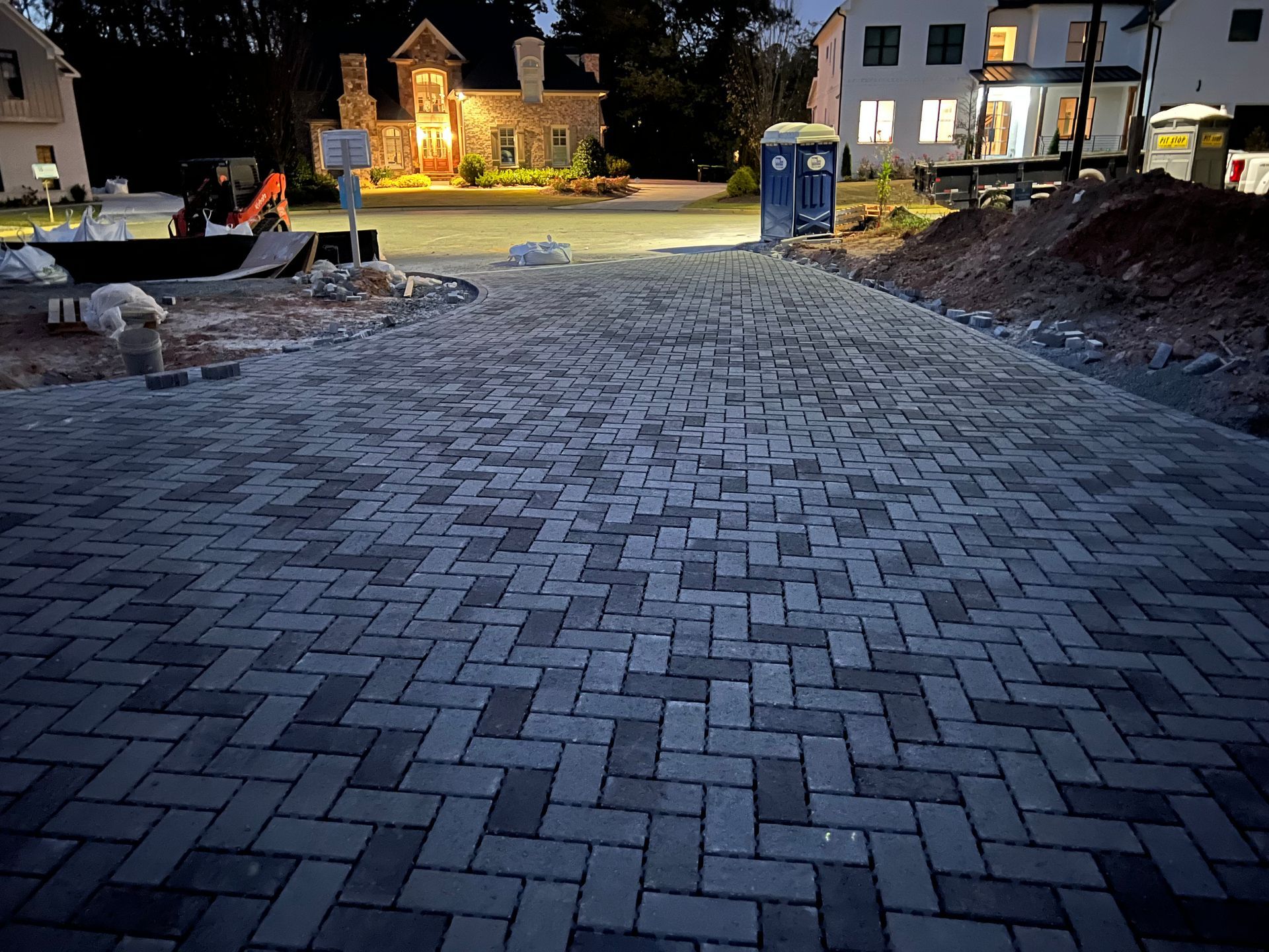 Paved brick driveway under construction at night, with a lit house in the background.