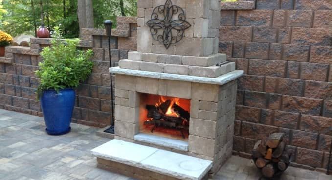Stone outdoor fireplace with a lit fire, set against a brick wall and patio planters