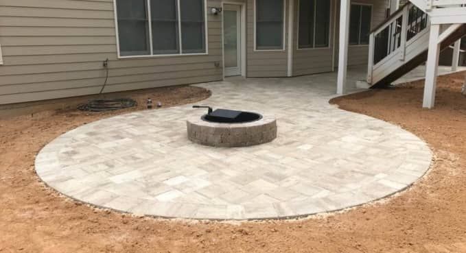 Backyard patio with a round paver fire pit and light stone seating area by a house