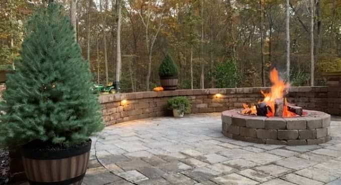 Backyard patio with stone fire pit burning beside potted evergreens and a low stone wall at dusk