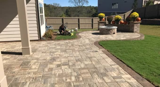 Stone patio and walkway beside a house, with a circular fire pit and flower planters next to a green lawn.