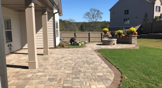 Backyard patio with stone pavers, beige house exterior, and a grassy lawn with flower planters.