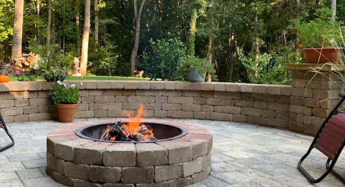 Stone patio fire pit with small flames, surrounded by chairs and potted plants in a wooded backyard