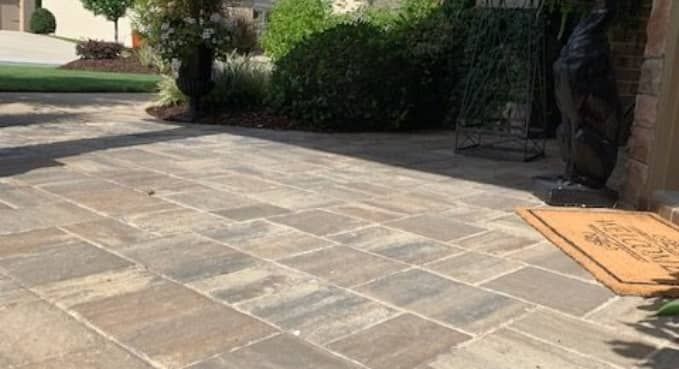 Stone patio with a tan doormat beside a hedge and driveway entrance