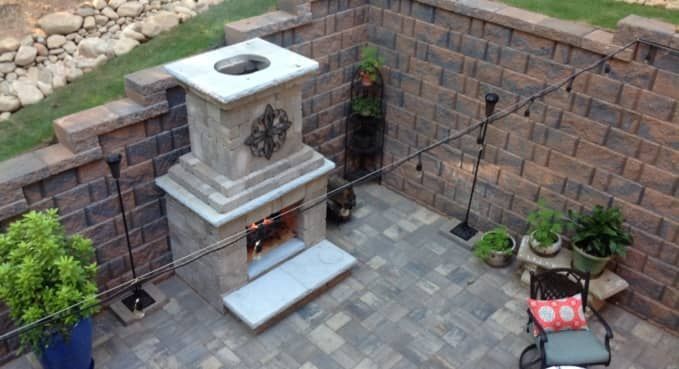 Small stone outdoor fireplace in a paved courtyard with brick walls and potted plants.