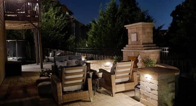 Patio with wicker chairs and dining table beside a lit brick fireplace at night