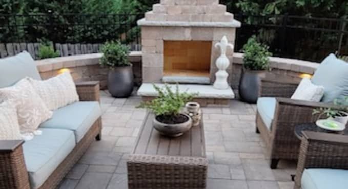 Outdoor patio seating area with wicker sofas, coffee table, and a stone fireplace surrounded by greenery