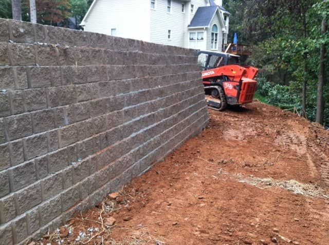 Construction site with orange skid steer leveling dirt beside a tall block retaining wall