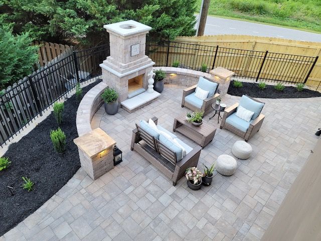 Patio with outdoor seating, stone fireplace, potted plants, and black fence beside a grassy yard.