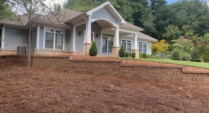 Single-story house with white columns and brick porch, set on a wooded hillside with mulch-covered front yard.