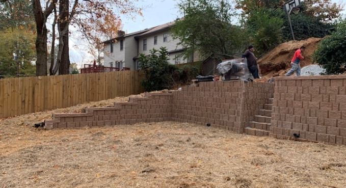 Backyard retaining wall under construction beside a fence, with workers and a house in the background