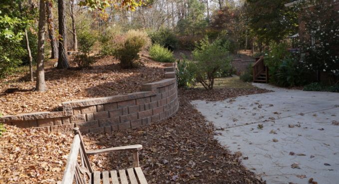 Leaf-covered backyard with stone retaining wall, bench, and paved patio under trees