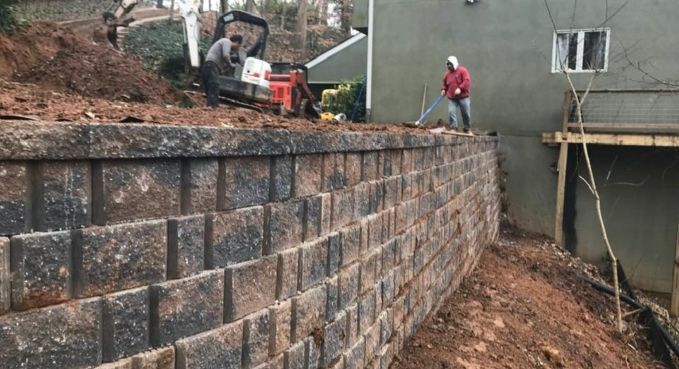 Workers building a stacked stone retaining wall beside a muddy excavation and house foundation