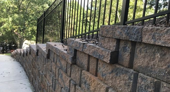Stone retaining wall beside a sidewalk, topped with black metal fencing and lined with trees.