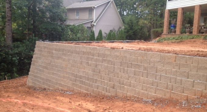 Tan retaining wall under construction beside a house, with reddish dirt and trees nearby