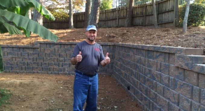 Man giving two thumbs up in front of a brick retaining wall in a wooded yard