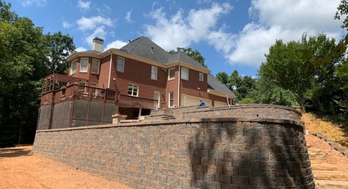 Large brick house on a raised stone wall under a blue sky with trees surrounding it.