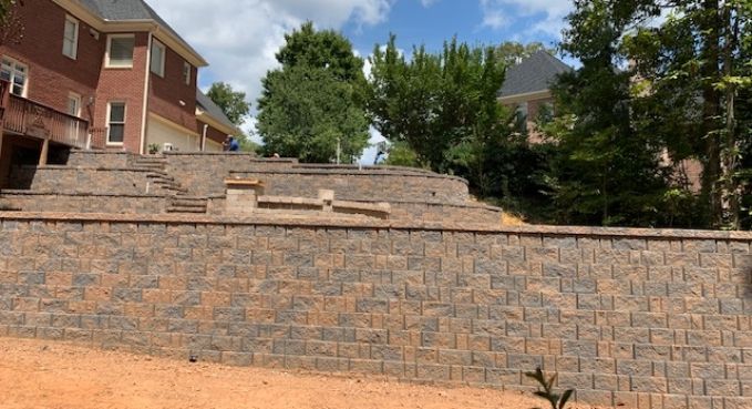 Stone retaining wall and terraced hillside beside brick houses and trees under a blue sky