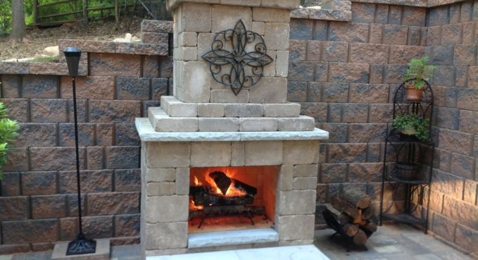 Stone outdoor fireplace with a lit fire, set against a brick retaining wall and patio seating area