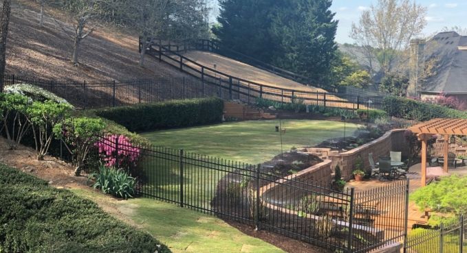 Landscaped backyard with a grassy slope, black fence, flower bed, and patio area under a pergola