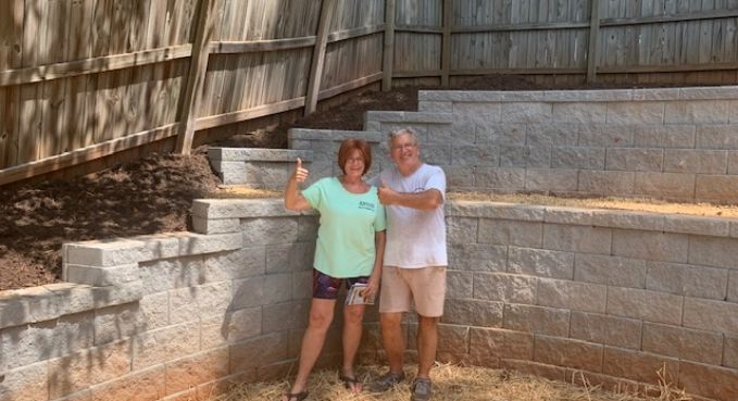 Two people standing together outdoors in front of a stone wall and wooden fence, smiling and posing.